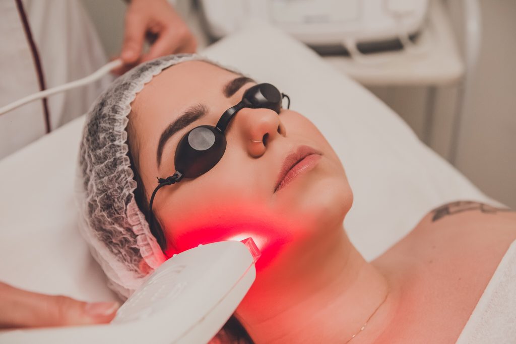 Pessoa deitada usando uma touca para proteger o cabelo e óculos de proteção sobre os olhos. Um profissional segura um dispositivo que emite uma luz vermelha, aplicando-o diretamente no rosto da pessoa. A cena ilustra um procedimento a laser, frequentemente utilizado em tratamentos estéticos e médicos, como rejuvenescimento da pele, remoção de manchas ou depilação.