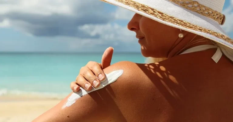 Uma pessoa aplica protetor solar no ombro enquanto aproveita um dia ensolarado na praia. Ela usa um chapéu branco de aba larga e um maiô claro. Ao fundo, o céu azul com algumas nuvens se encontra com as águas turquesa do oceano. A cena destaca a importância da proteção solar durante atividades ao ar livre.