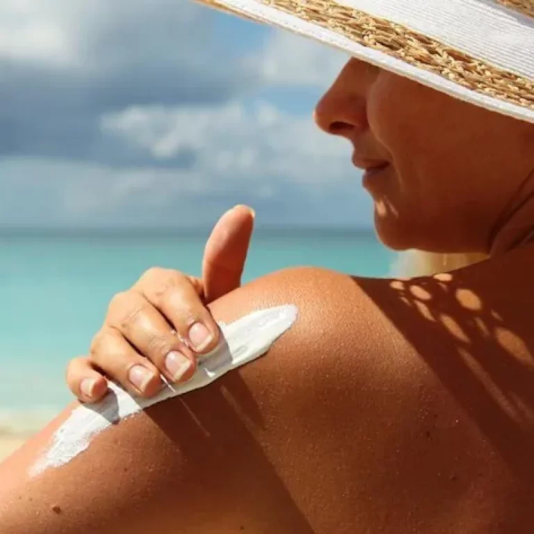 Uma pessoa aplica protetor solar no ombro enquanto aproveita um dia ensolarado na praia. Ela usa um chapéu branco de aba larga e um maiô claro. Ao fundo, o céu azul com algumas nuvens se encontra com as águas turquesa do oceano. A cena destaca a importância da proteção solar durante atividades ao ar livre.