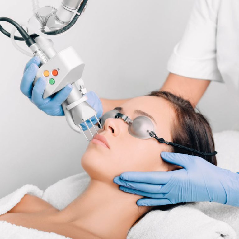 Profissional de saúde vestindo uniforme branco e luvas azuis, segurando um dispositivo médico conectado a uma máquina maior por meio de cabos. O paciente está deitado, com uma toalha branca cobrindo parte do corpo, enquanto o profissional aplica o dispositivo na região do pescoço. A presença de botões coloridos no aparelho sugere um procedimento técnico, possivelmente relacionado a tratamentos estéticos ou terapias médicas.